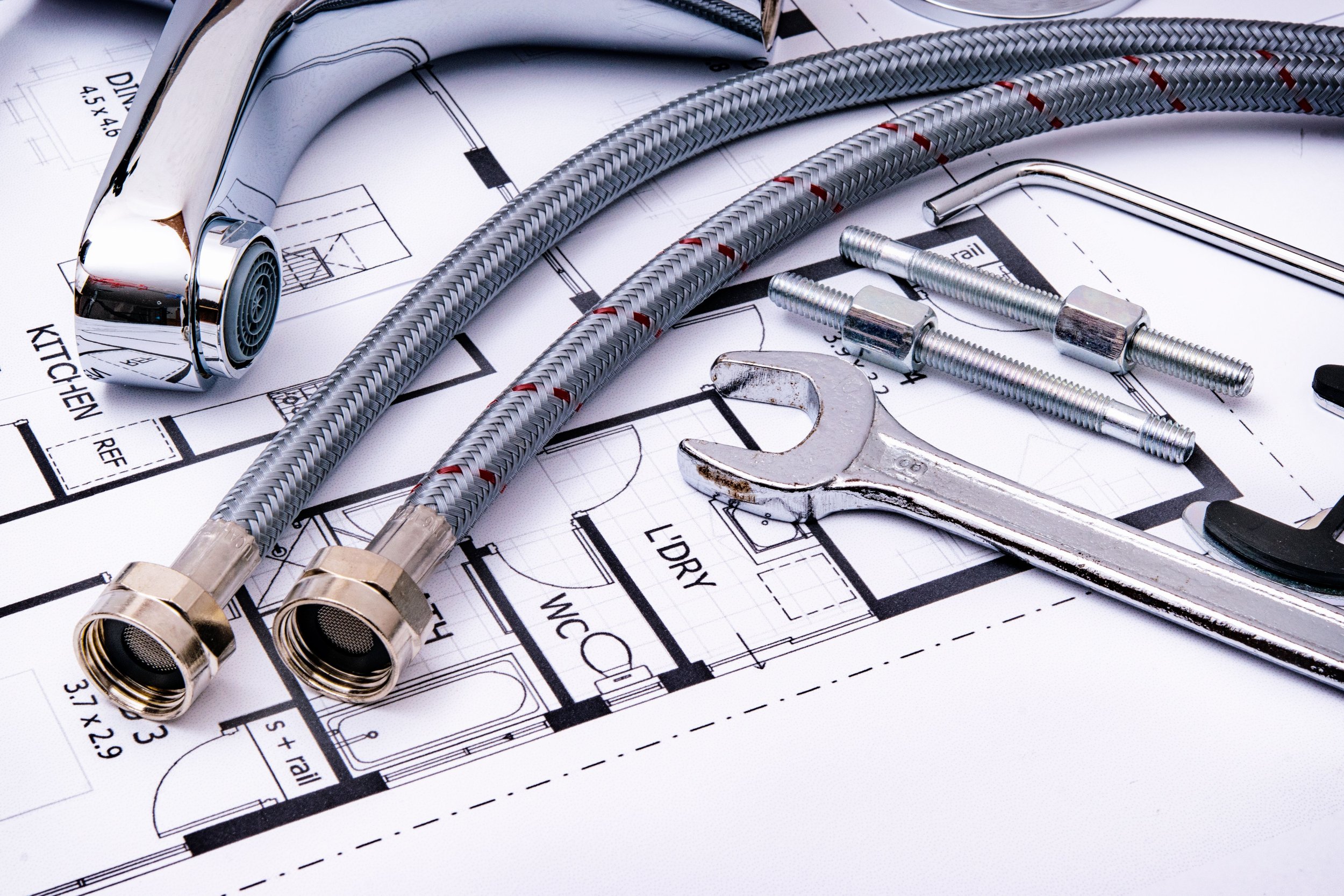 Various plumbing tools and pipes laid out on top of blueprints, including braided hoses, a wrench, screws, and a chrome drain component.