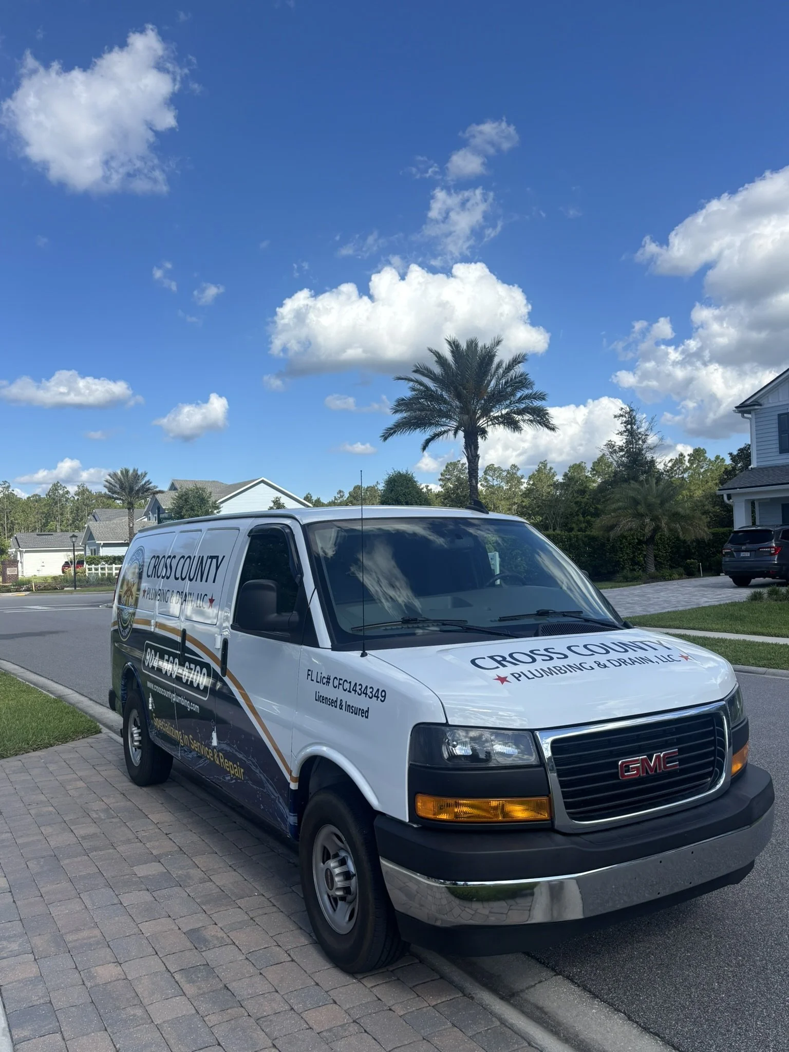 GMC van belonging to Cross County Plumbing & Drain LLC parked on a residential driveway with houses, trees, and a palm tree under a partly cloudy sky.