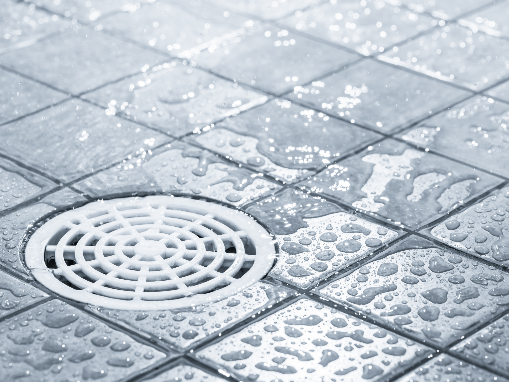 Close-up of a wet outdoor drain with a round plastic grate, surrounded by wet tiles and water droplets.