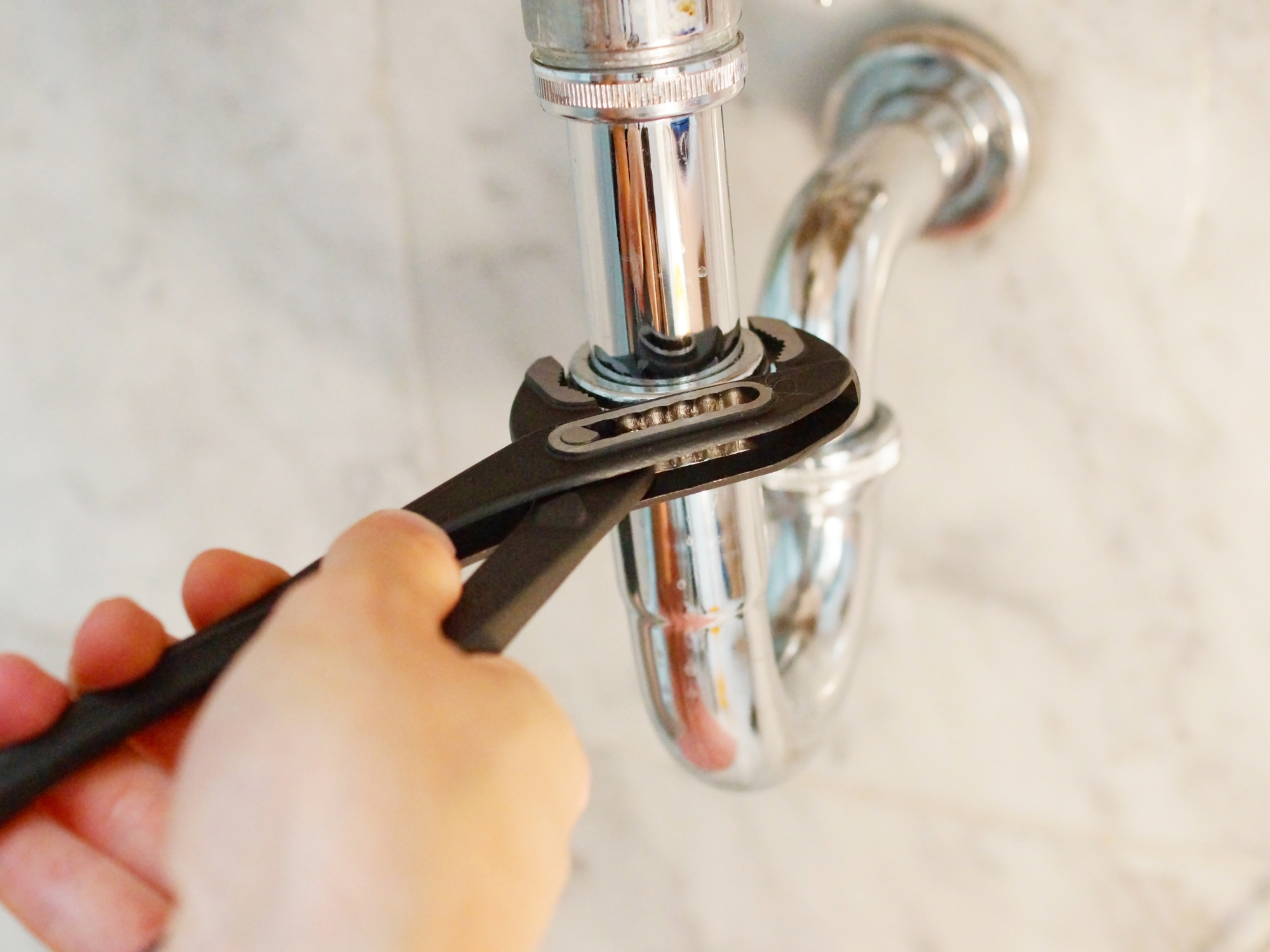 Person using pliers to work on plumbing under a sink with shiny chrome pipes.