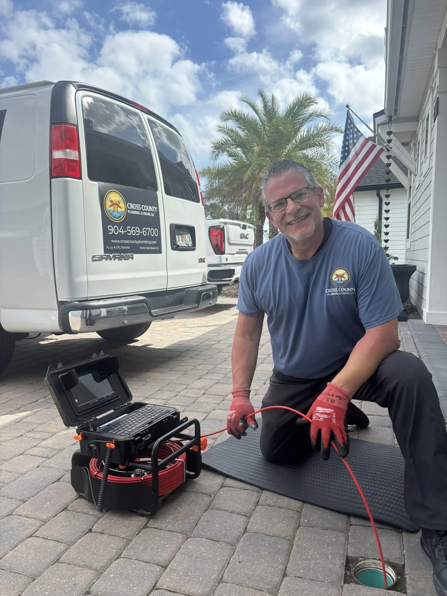 A man wearing glasses and a blue shirt with a plumbing company logo kneels on a paved driveway next to an open hole with a red cable running into it. There is a toolbox with testing equipment and a laptop nearby. Behind him, a white van with the same logo and phone number is parked, with palm trees and a house in the background, under a partly cloudy sky.