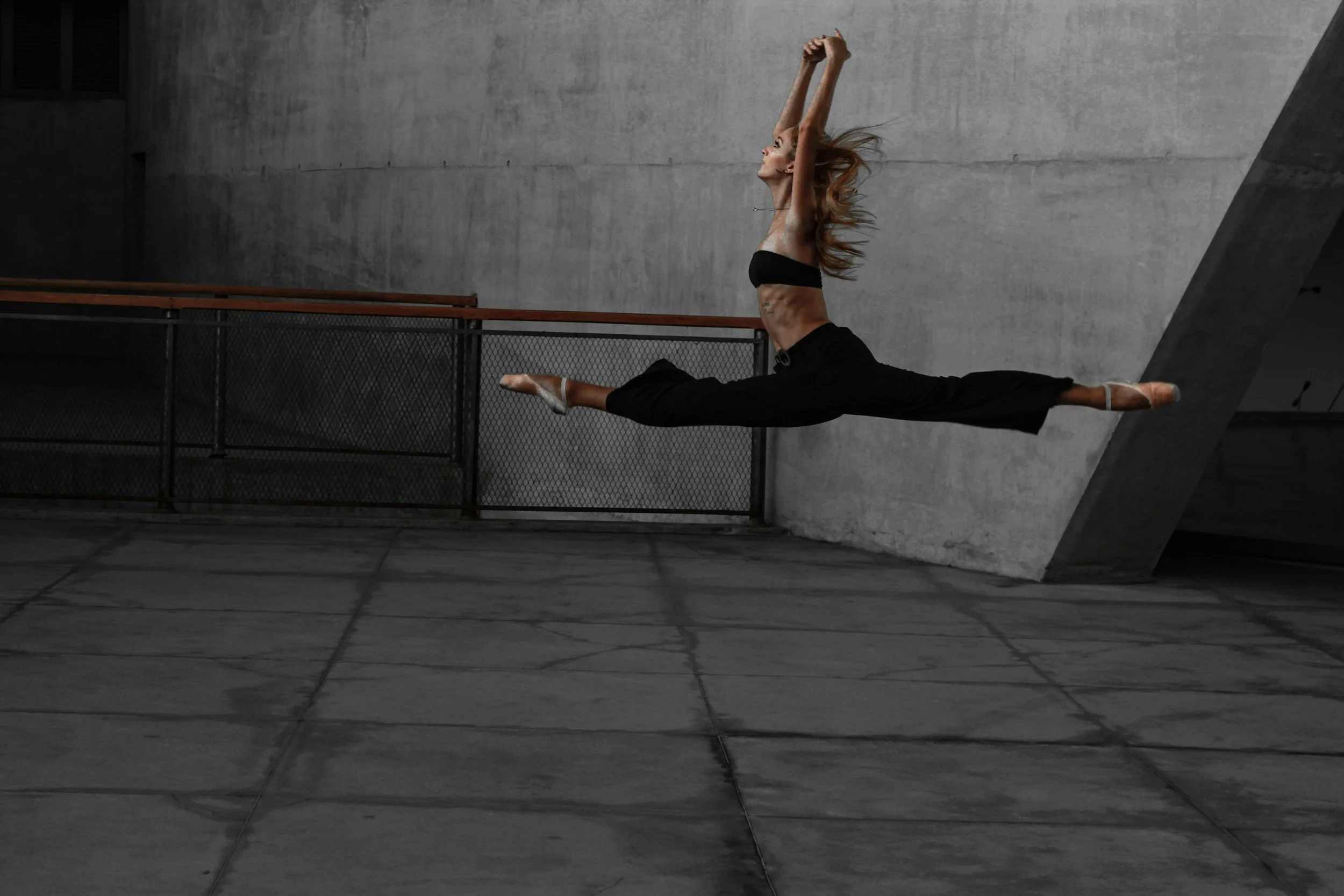 A woman performing a split dance move against a concrete wall, with arms stretched upward and flowing hair.