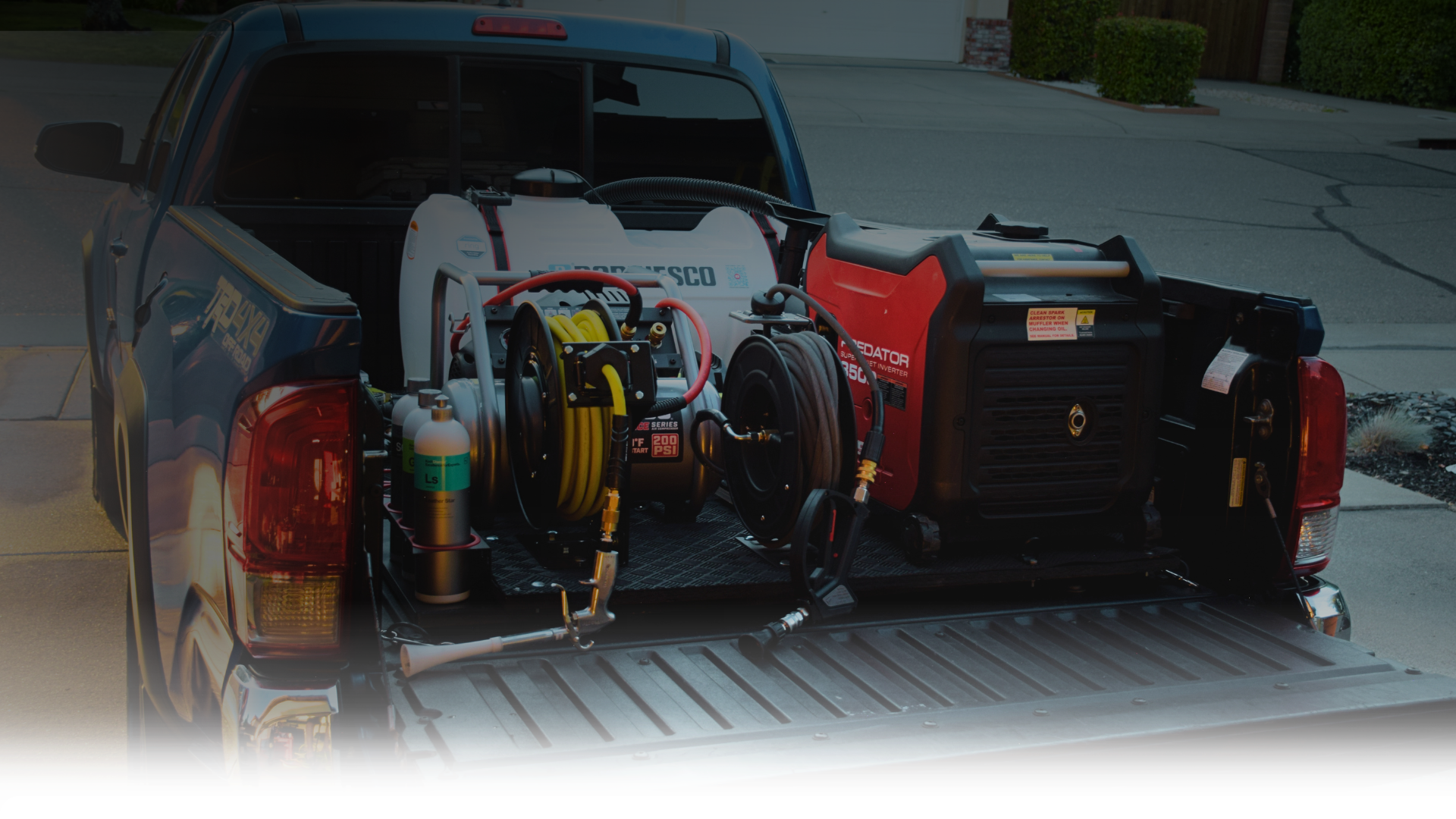 The back of a pickup truck containing various equipment including a red generator, hoses, and a white tank.