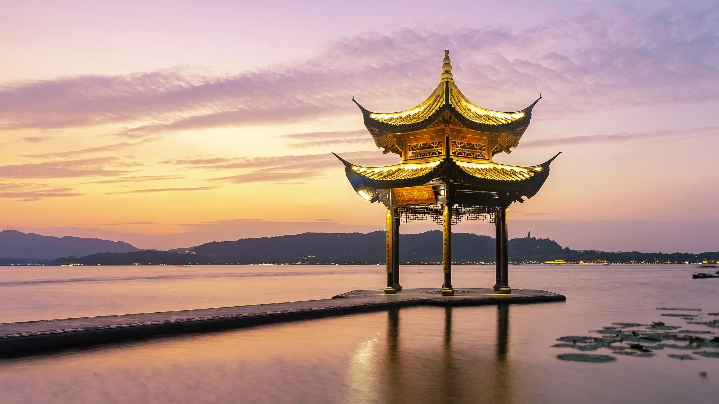 A traditional Chinese pavilion illuminated at sunset, situated on water with a mountain and distant city lights in the background.