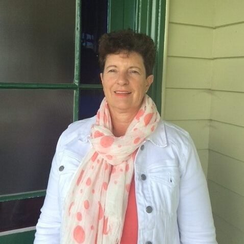 A woman with short curly hair wearing a white jacket and pink polka dot scarf, standing outside near a green door and beige siding.