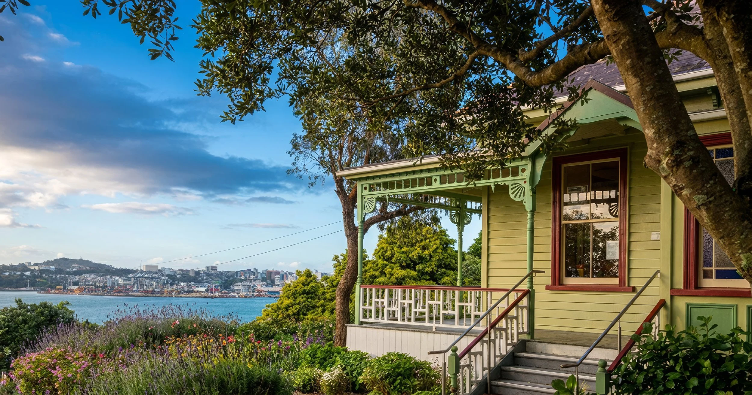 A colorful house with green and yellow walls, a porch with decorative trim,, and a view of the city skyline across a body of water, framed by a large tree and blooming flowers.