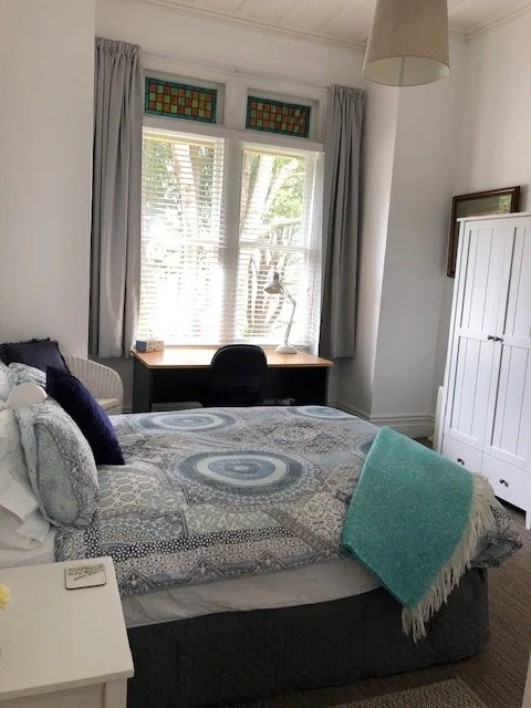 Gray bedroom with a bed, navy pillows, light blue throw blanket, window with blinds and stained glass, wooden desk and black chair, white cabinet, and beige hanging lamp.