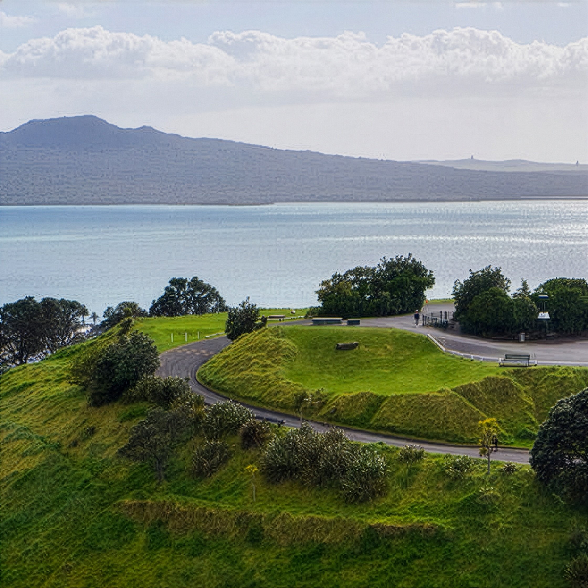A scenic view of a hill with lush green grass and trees, a winding path, and a body of water in the background under a cloudy sky.