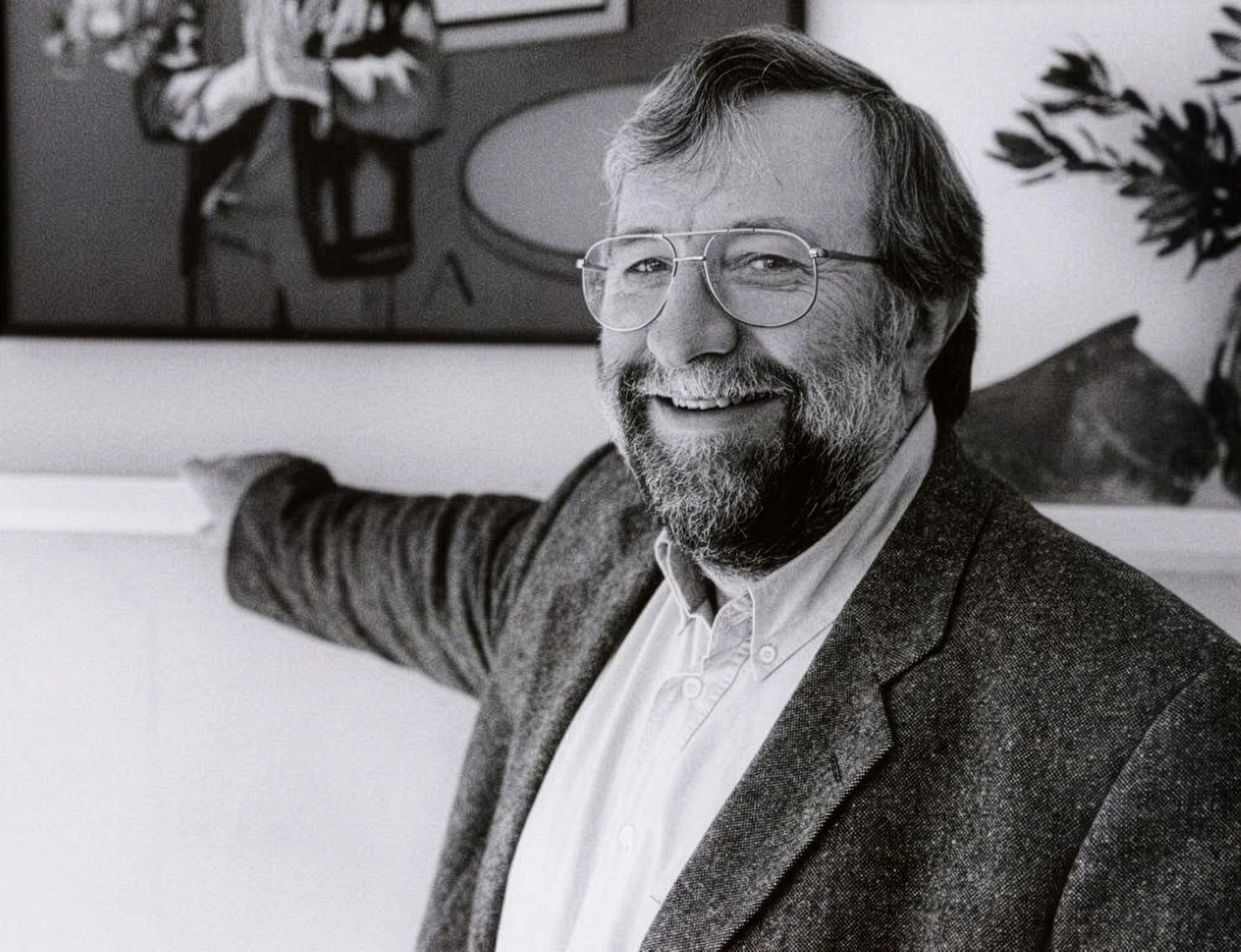 Black and white photo of a smiling man with glasses, beard, and mustache, wearing a blazer over a light shirt, sitting with his right arm extended, in a room with art and plants.