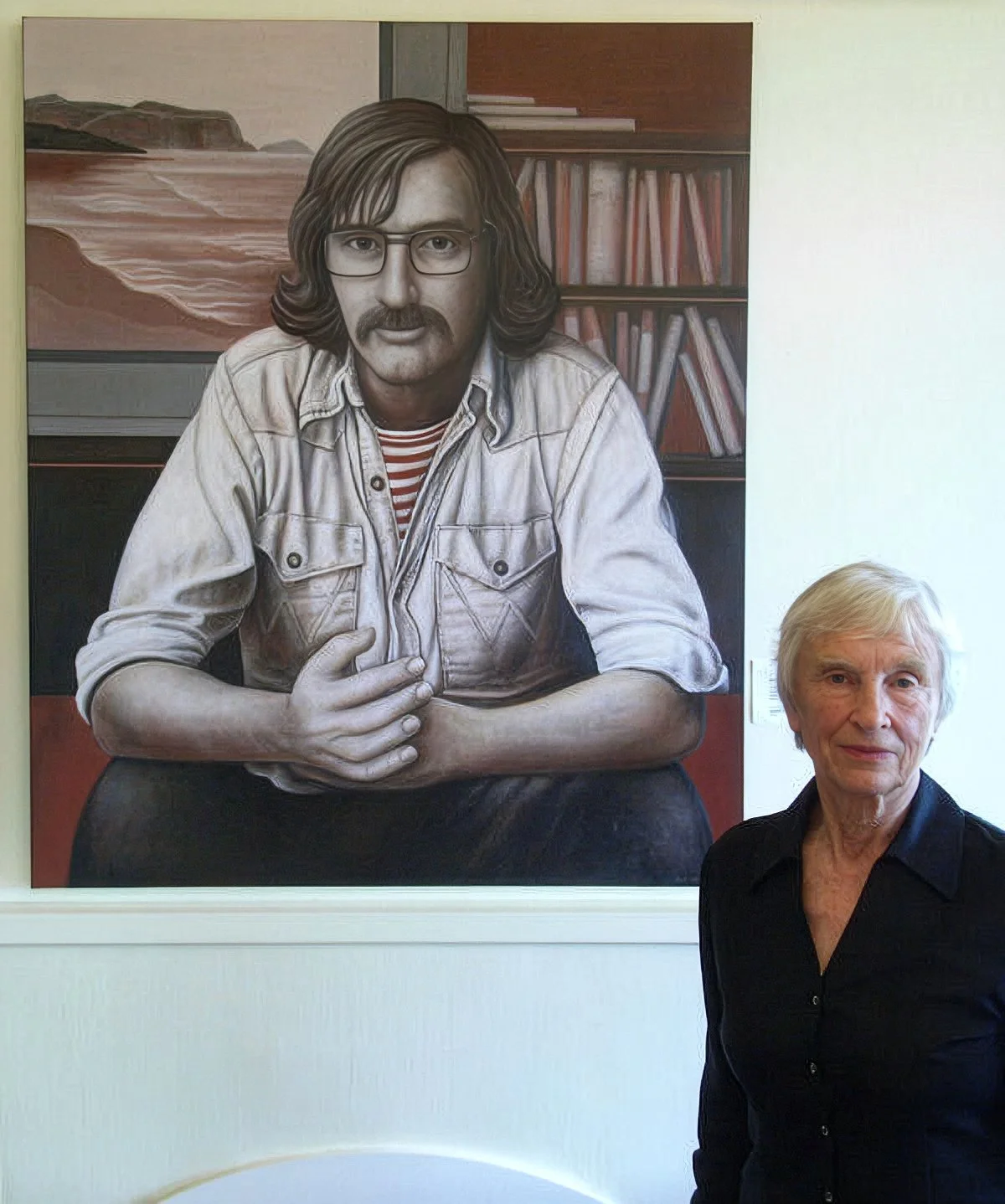 A woman standing next to a large portrait of a man with long hair, glasses, a mustache, and a beard. The man in the painting is wearing a striped shirt and a light-colored jacket, sitting with his arms crossed on his lap. The background of the portrait features a bookshelf with books.