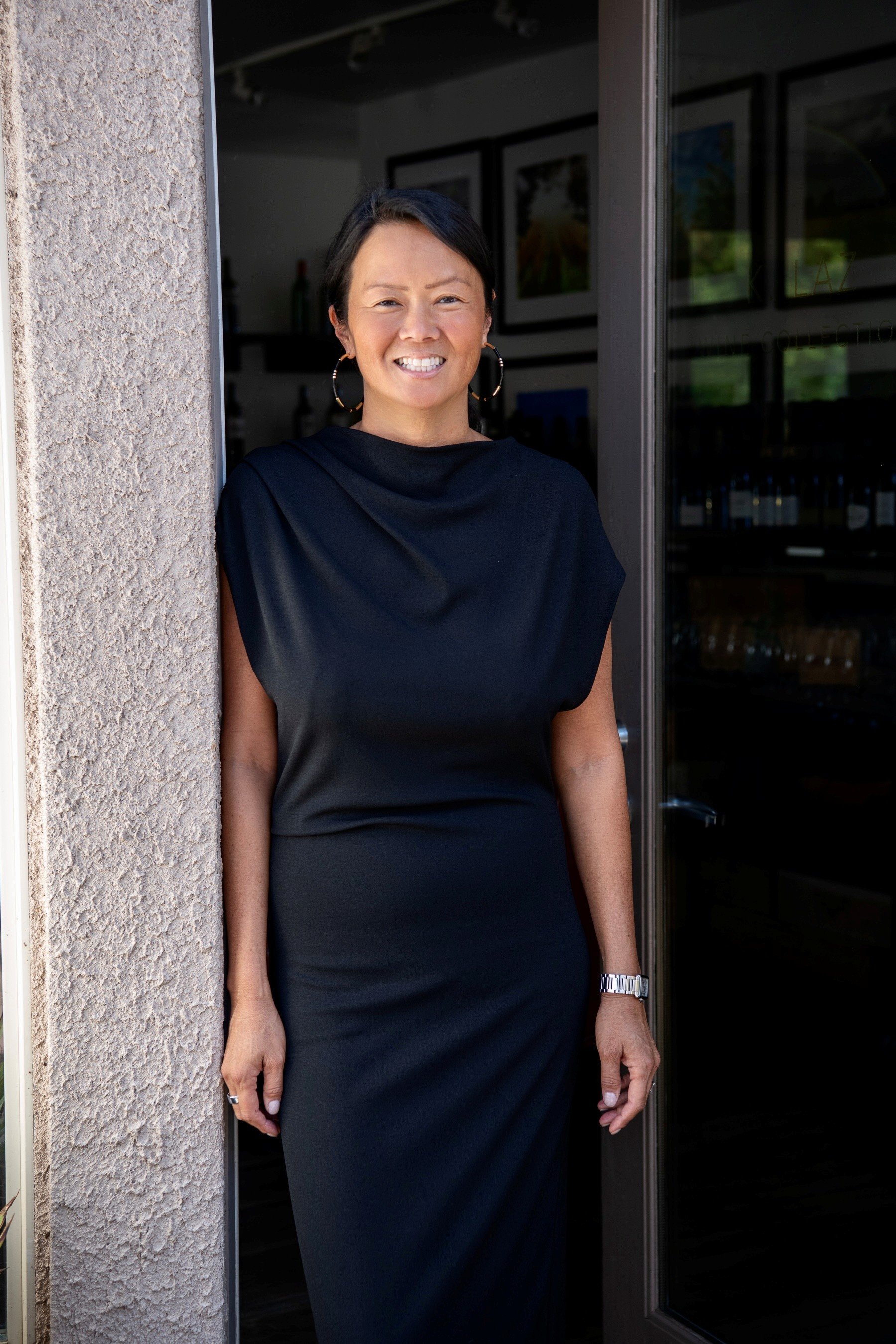 Founder Kerrin Laz with short black hair stands in an open doorway, wearing a black dress with a high neckline, large hoop earrings, a silver watch on her left wrist, and a ring on her right hand. She is leaning against a textured beige wall.