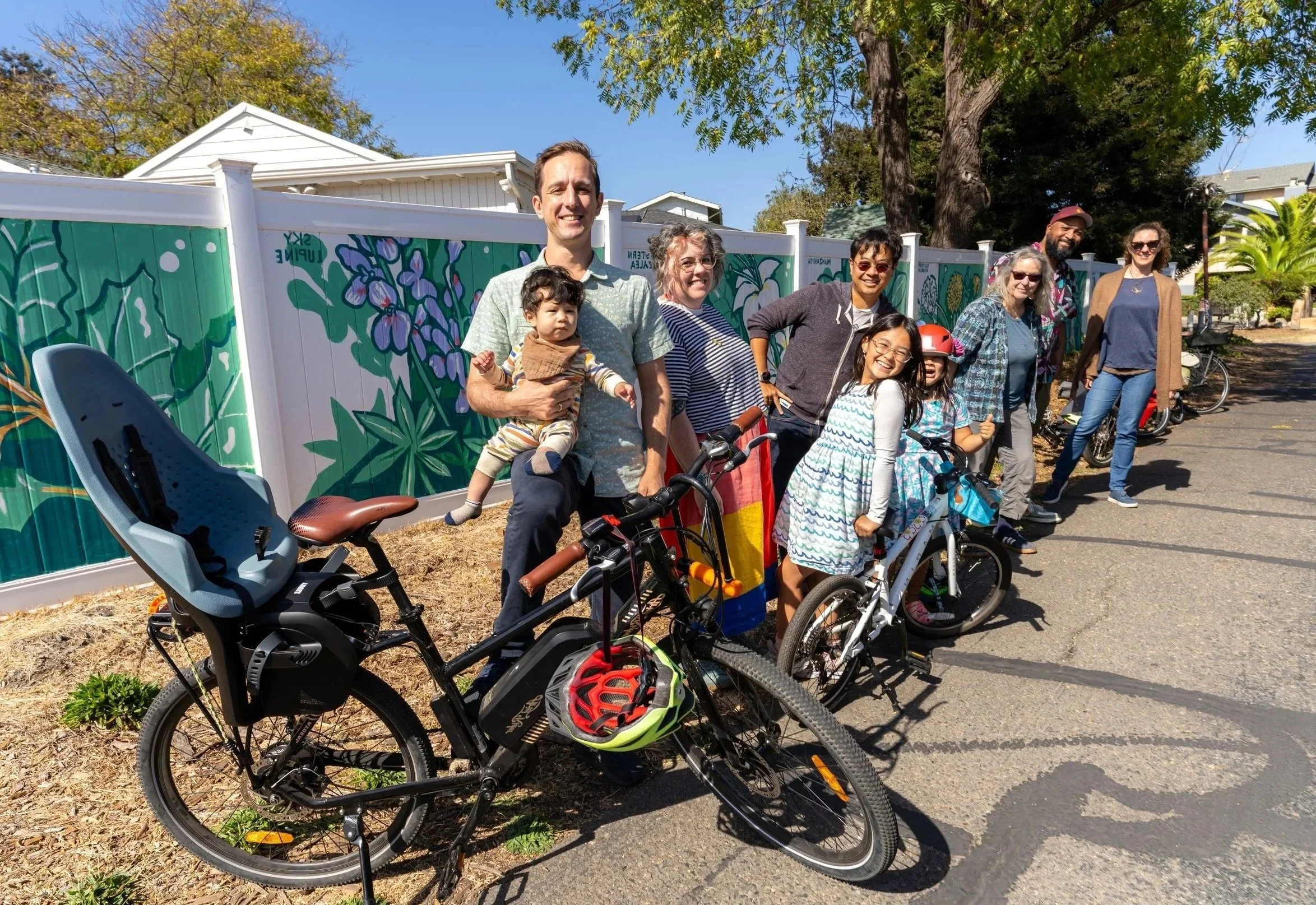 Group of people smiling and standing with bicycles outdoors on a sunny day, with trees and a painted fence in the background.