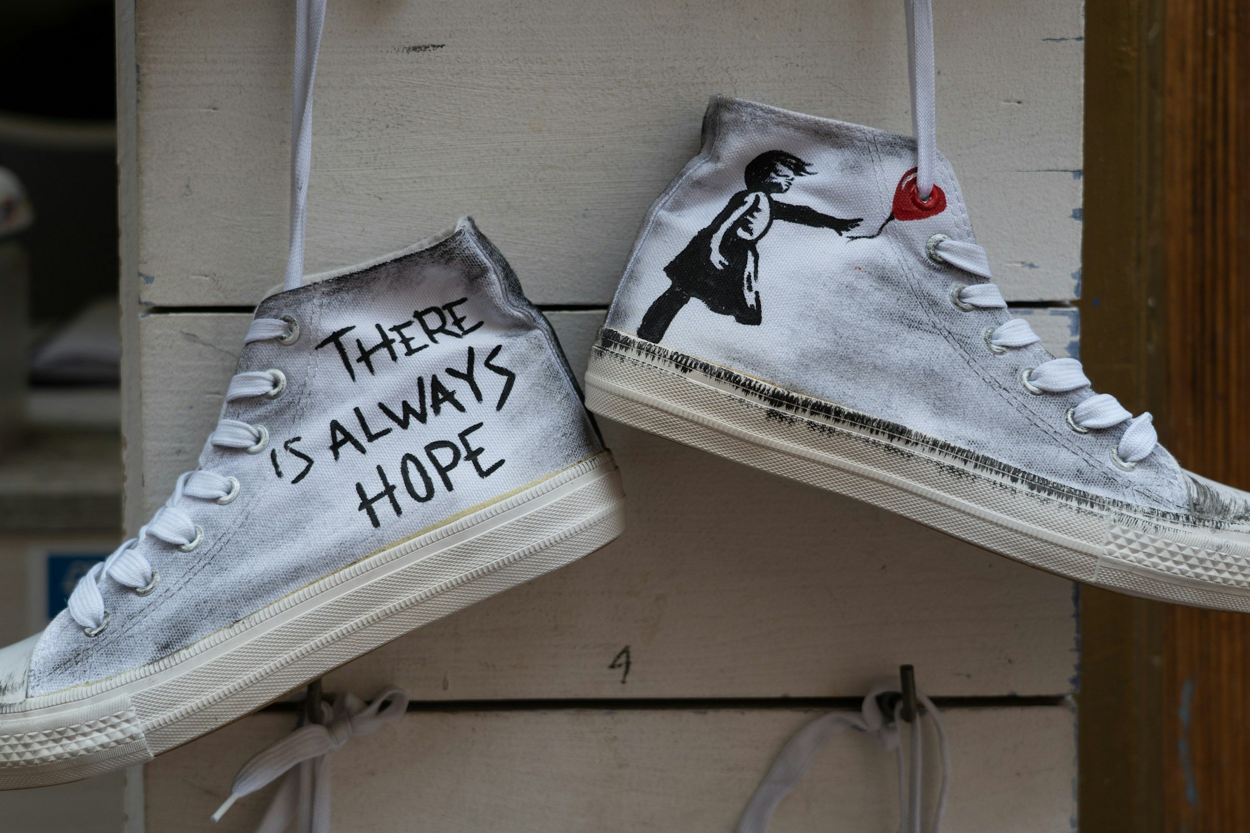 A pair of white canvas sneakers with painted designs and text are hanging on a wooden display board. One shoe has the phrase 'There is always hope' written across it, and the other shoe features a silhouette of a girl reaching out to a red heart.