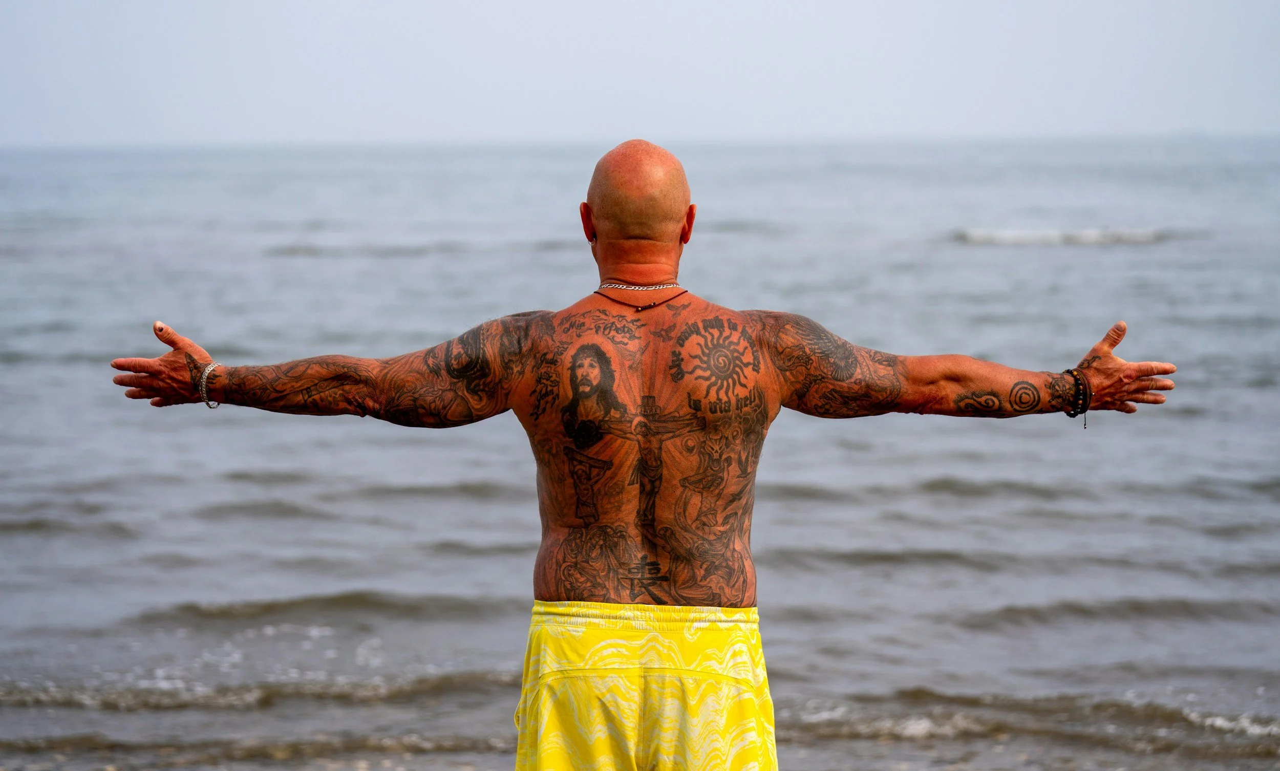 A bald, tattooed man wearing yellow shorts standing on a beach with arms outstretched towards the ocean.