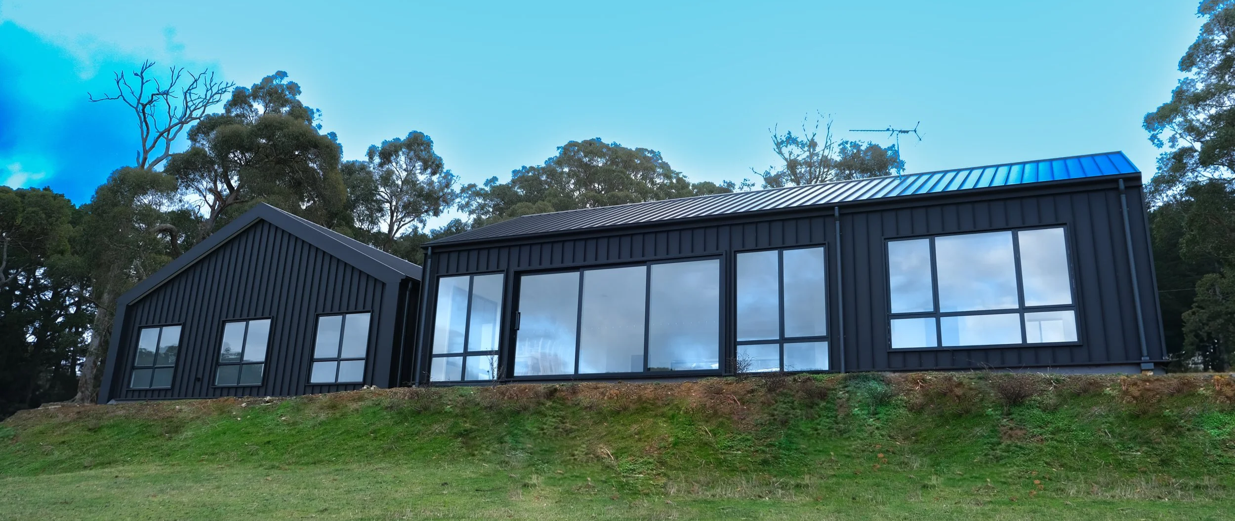 Modern black house with large windows, situated on a grassy hill with trees in the background under a blue sky.