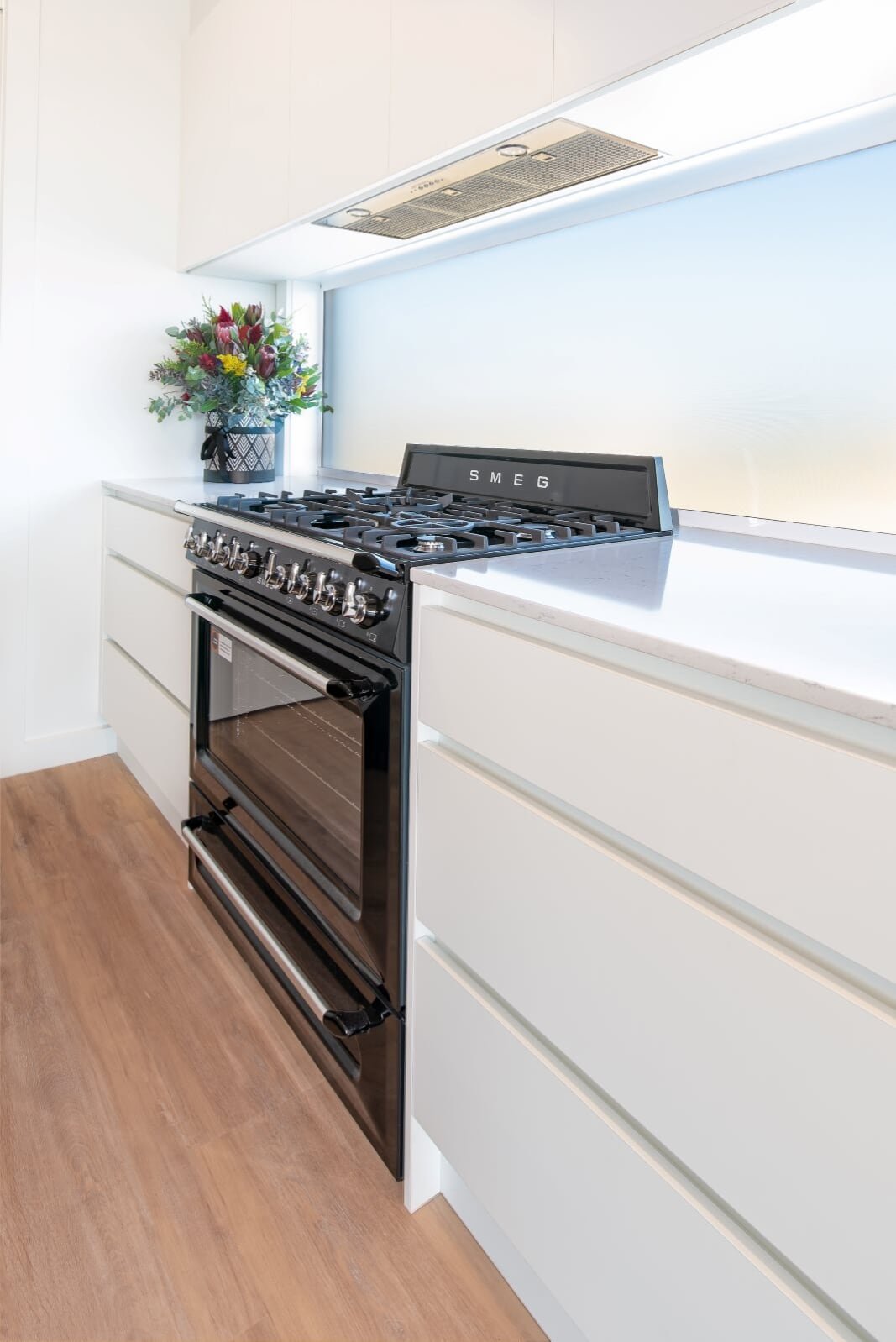 Modern kitchen with white cabinets, a black SMEG oven, a gas stove, a bouquet of flowers in a black decorative vase, and wooden flooring.