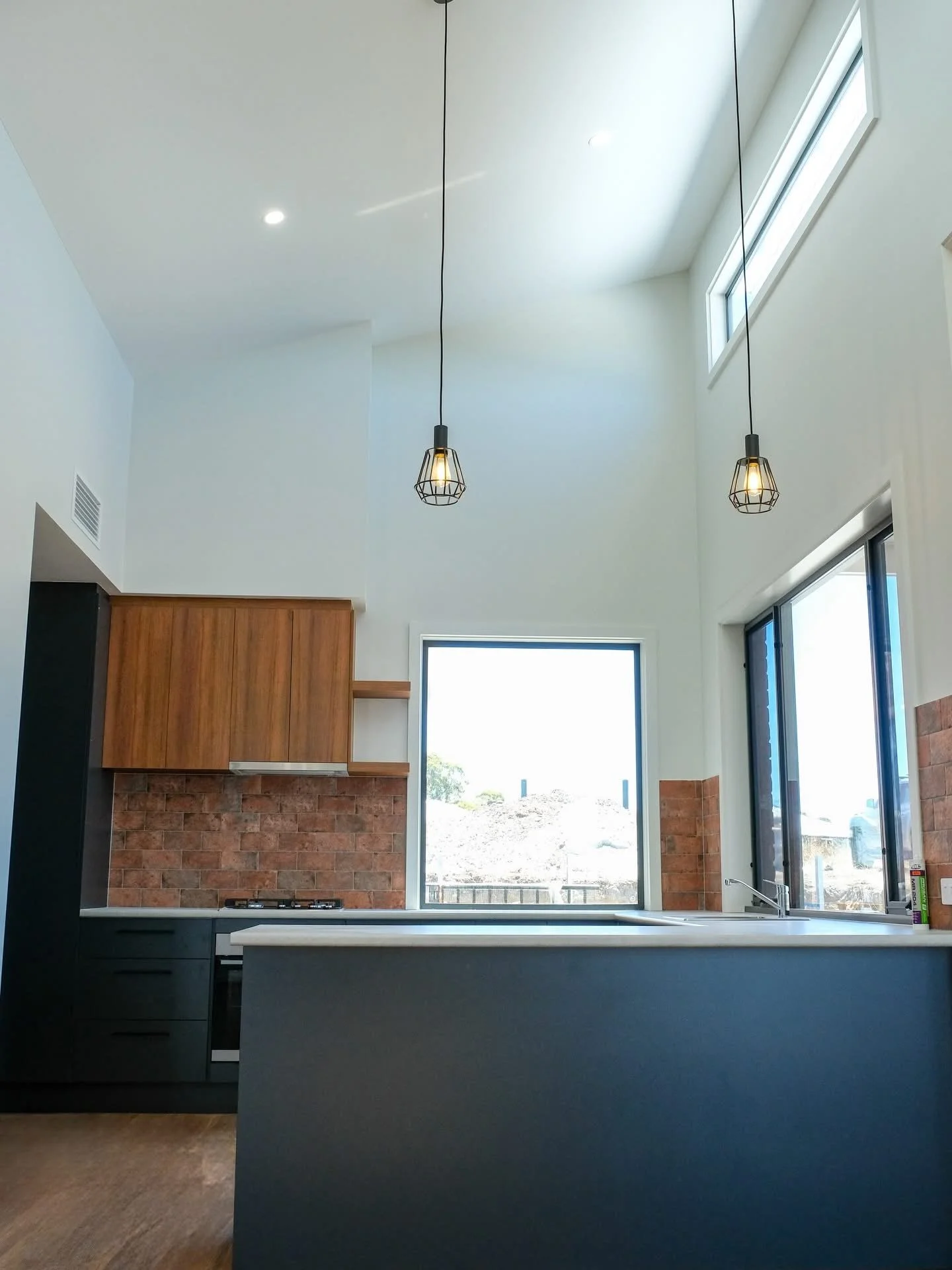 Modern kitchen with white walls, large windows, wooden cabinets, exposed brick backsplash, and black and white countertops, illuminated by pendant lights.