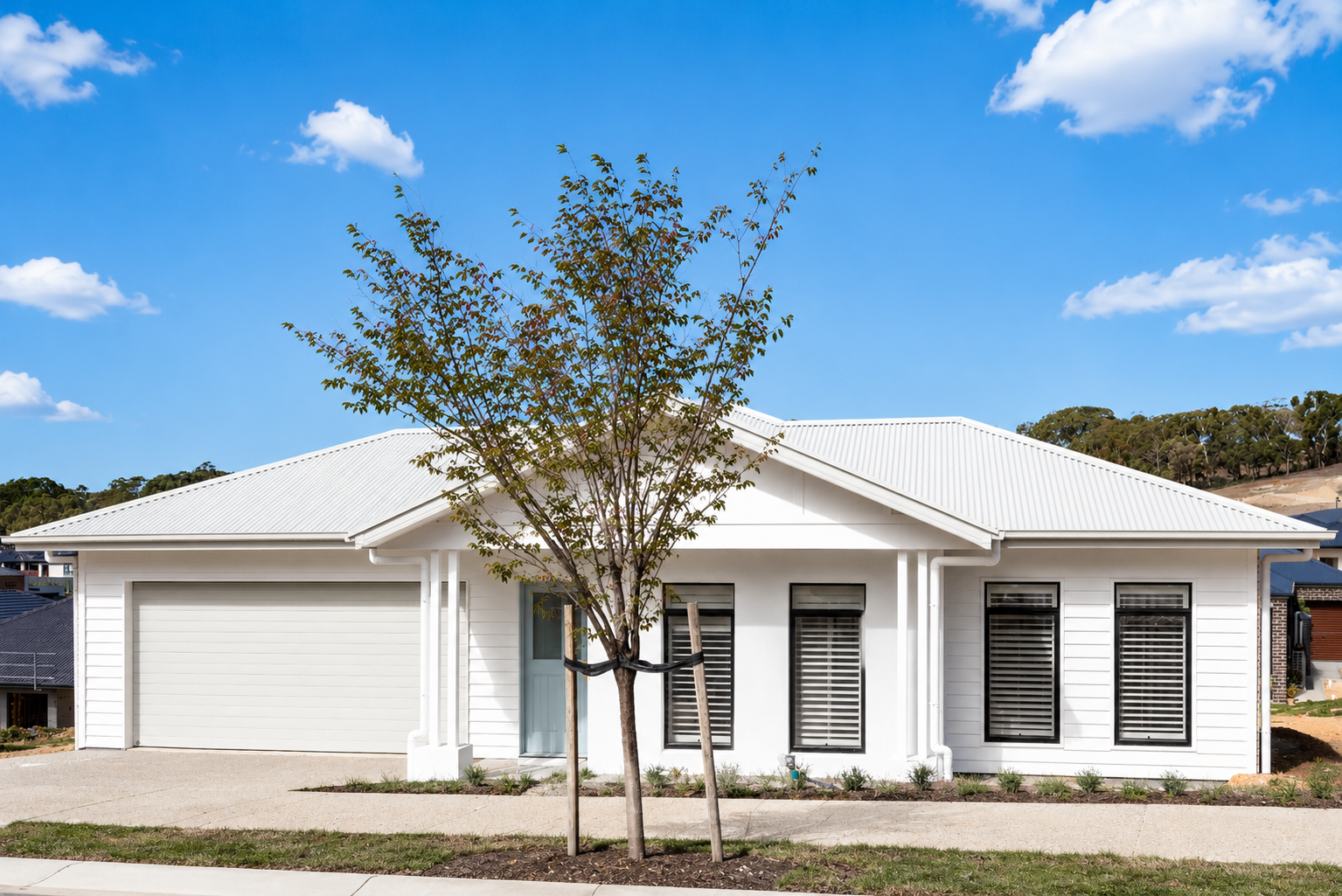 White modern house with a metal roof, three windows with black shutters, a garage door, and a sidewalk with a leaning tree in front.