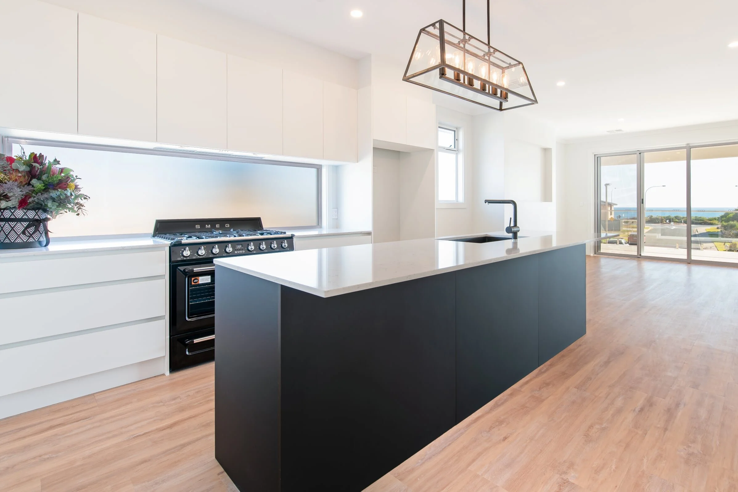 Modern kitchen with white cabinets, black island, wooden flooring, and sliding glass door overlooking outdoors. There is a floral arrangement on a white counter, a black oven, and a large pendant light fixture overhead.