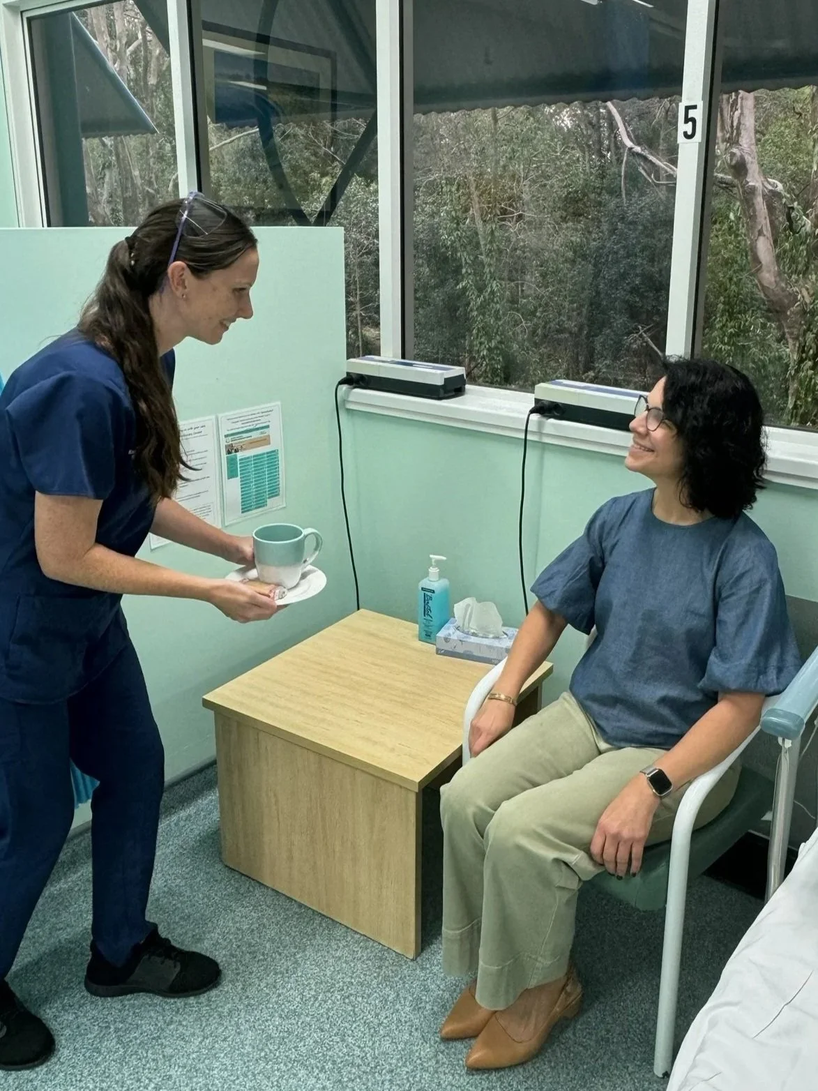 A woman in medical scrubs is handing a mug to a seated woman in a medical gown in a clinic room. The room has large windows, a small table with hand sanitizer and tissues, and medical equipment.