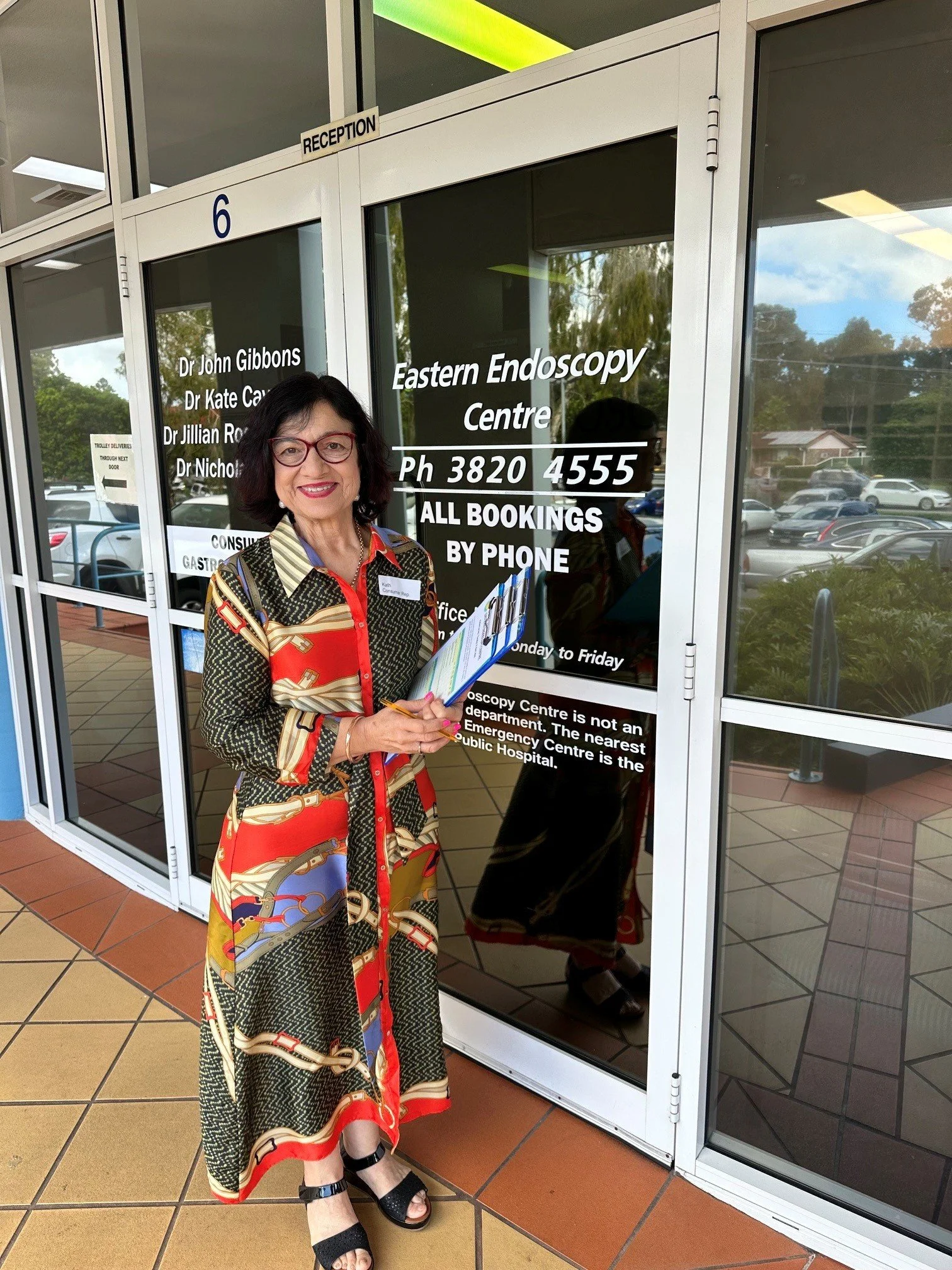 A woman standing outside the Eastern Endoscopy Centre in front of glass doors with information and contact details printed on them. She is holding documents and wearing a colorful patterned dress with black sandals, smiling at the camera.