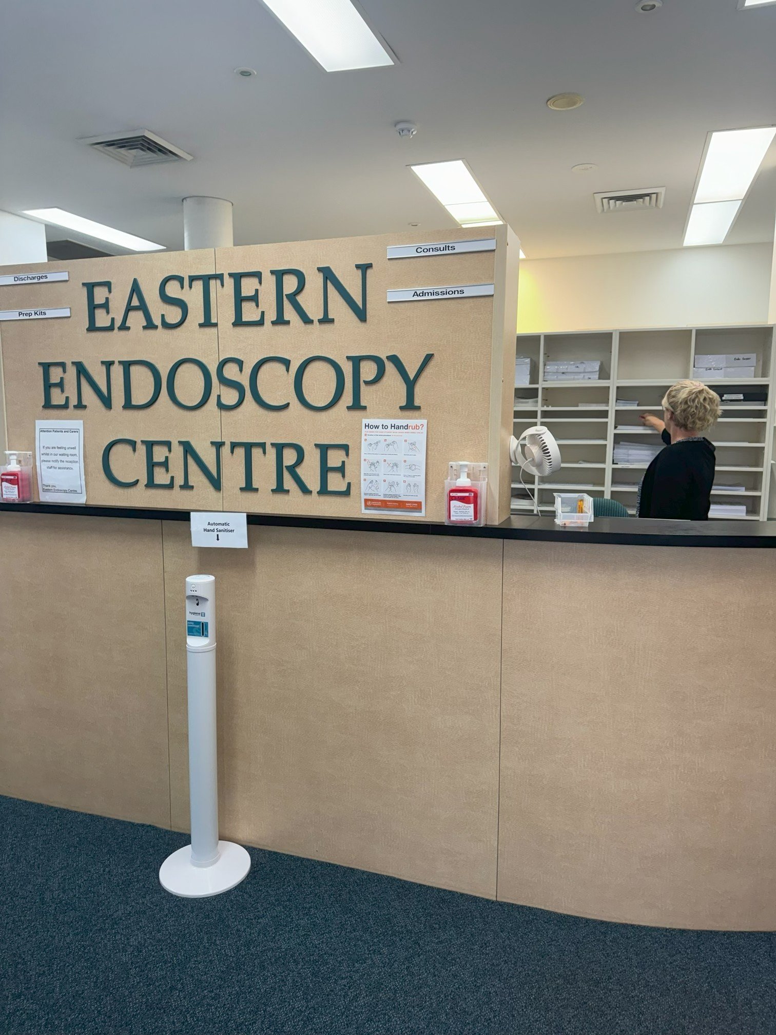 Reception desk at Eastern Endoscopy Centre with a large sign displaying the center's name, a hand sanitizer dispenser, and a person organizing files on shelves in the background.