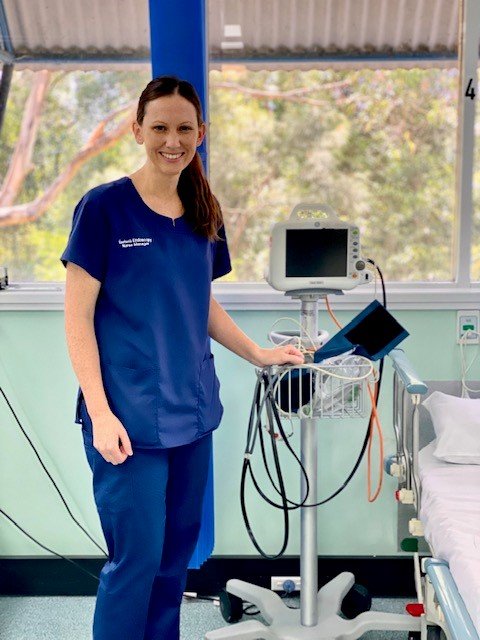 A female nurse in blue scrubs standing in a hospital room next to a medical monitor and equipment, smiling at the camera.