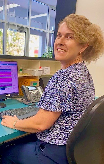 A woman with curly blonde hair smiling at the camera while sitting at an office desk with a computer monitor, phone, and office supplies.