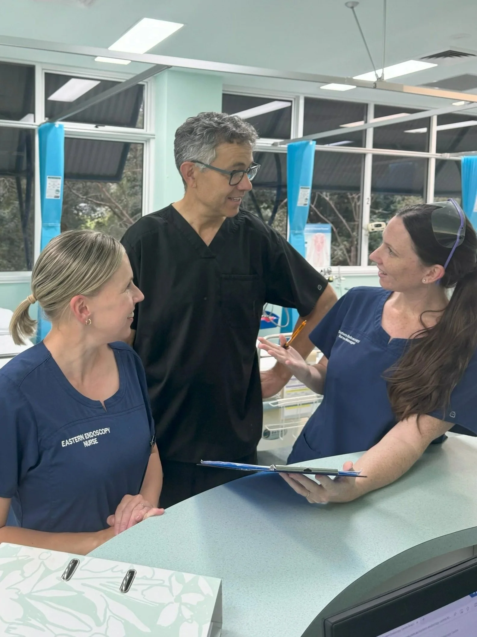 Medical professionals in scrubs having a discussion in a clinic or hospital room, with windows and medical equipment in the background.