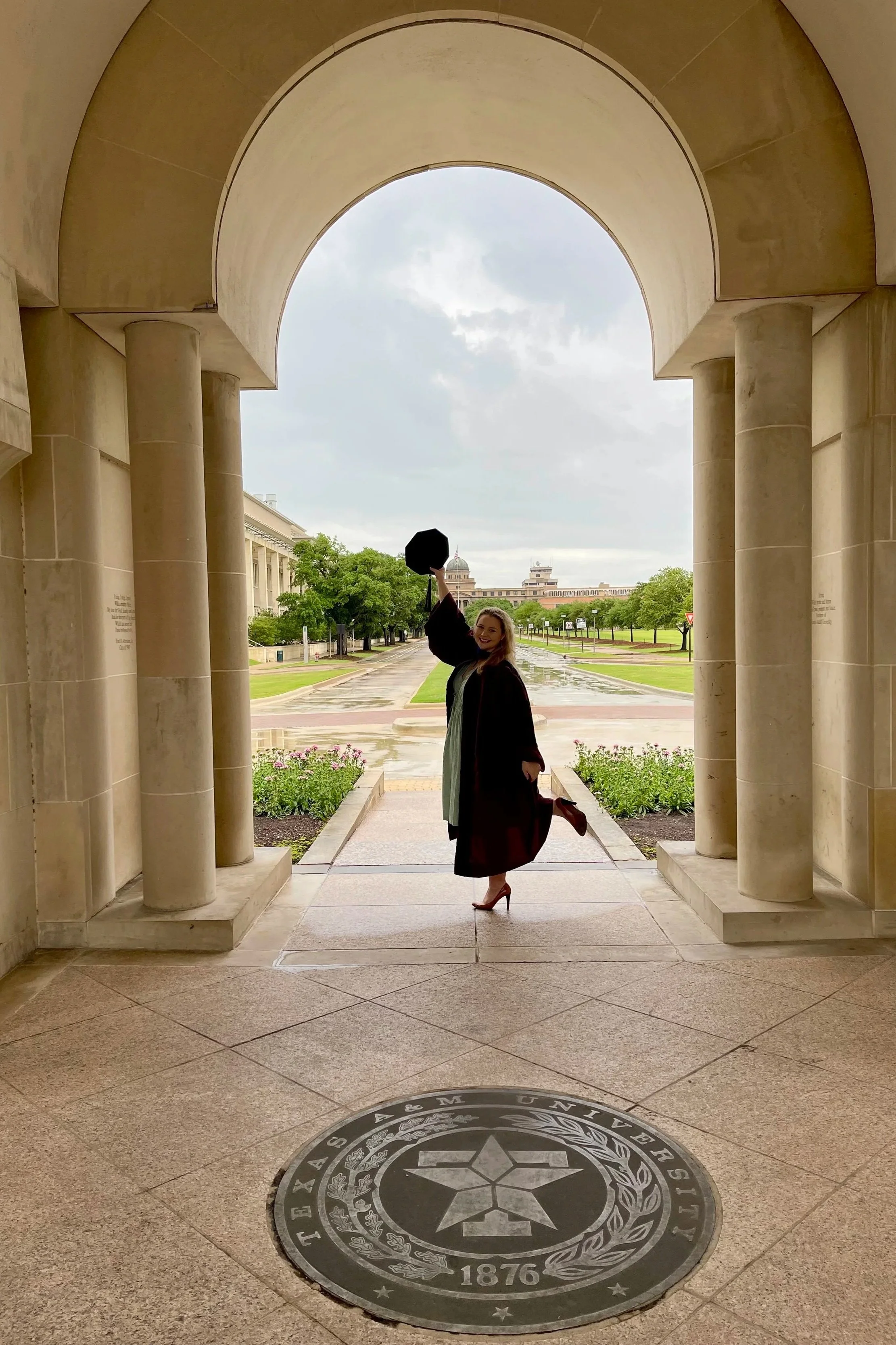 A woman in a graduation cap and gown standing under a stone archway, holding her cap in the air and smiling. The background shows a campus with trees, pathways, and buildings, with overcast sky.