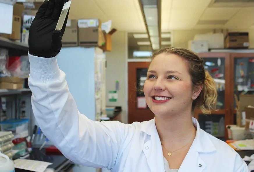 A young female scientist in a laboratory taking a selfie with her smartphone. She is smiling and wearing a white lab coat and black gloves.