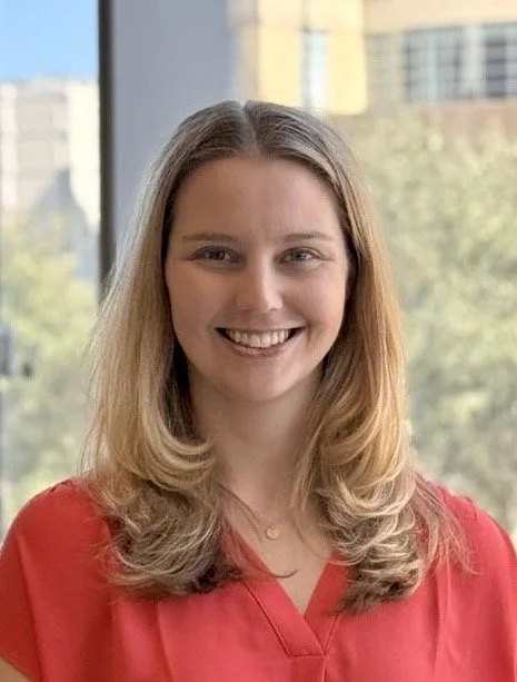 A young woman with long blonde hair, wearing a red top, smiling in front of a large window with a cityscape in the background.