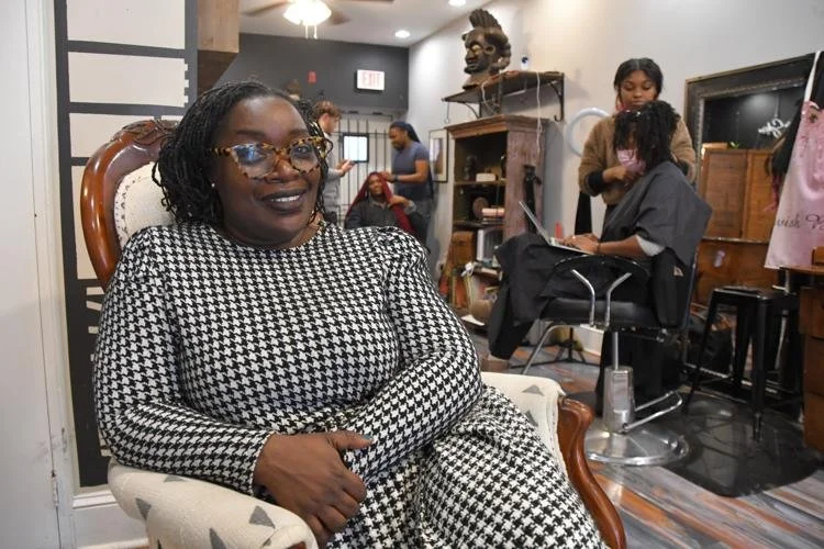 Woman sitting in a salon chair wearing a black and white houndstooth dress, smiling, with hair styled and wearing glasses; people in the background getting hair treatments.