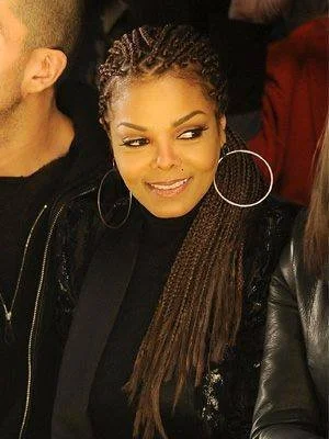 A woman with long, braided hair, wearing large hoop earrings and a black jacket, looking to her left with a smile.