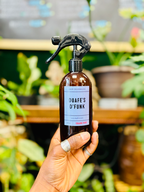 Hand holding a brown spray bottle with a black nozzle labeled 'DUAFE'S D'FUNK' against a background of potted plants and a gardening area.