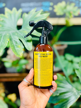 A hand holding a spray bottle labeled 'BAREL'S FLAX SEED & RICE WATER' in front of green leafy plants.