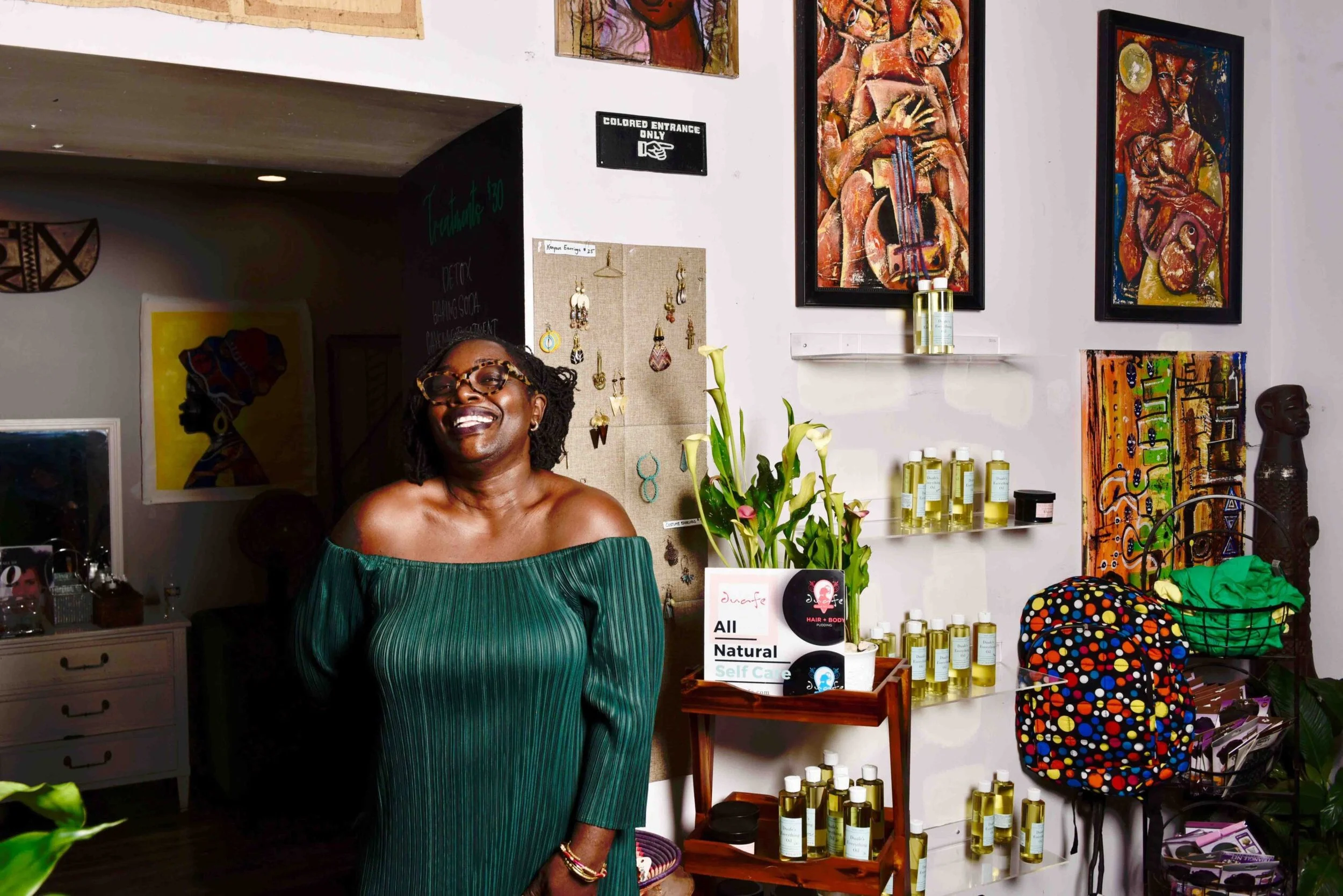 A woman with glasses and braided hair, smiling, stands in a colorful art gallery or shop. Behind her are vibrant paintings, artwork, and jewelry displayed on the white wall, as well as shelves with bottles and containers. The scene suggests a cozy, artistic setting.