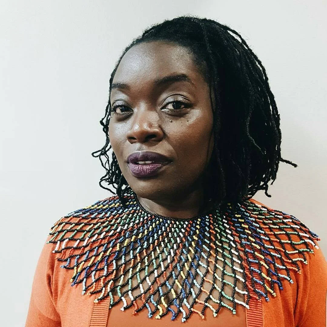 Portrait of a Black woman with black dreadlocks wearing an orange top and a colorful beaded necklace.