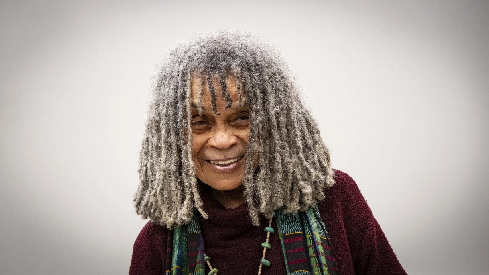 A smiling older woman with gray dreadlocks wearing a maroon sweater and colorful beaded necklaces, standing against a plain gray background.