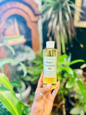 A hand holding a bottle of Duafe's Everything Oil in a lush green garden or indoor plant setting.
