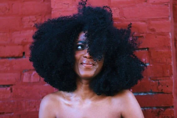 A woman with large, curly black hair smiling and looking to the side in front of a red brick wall.