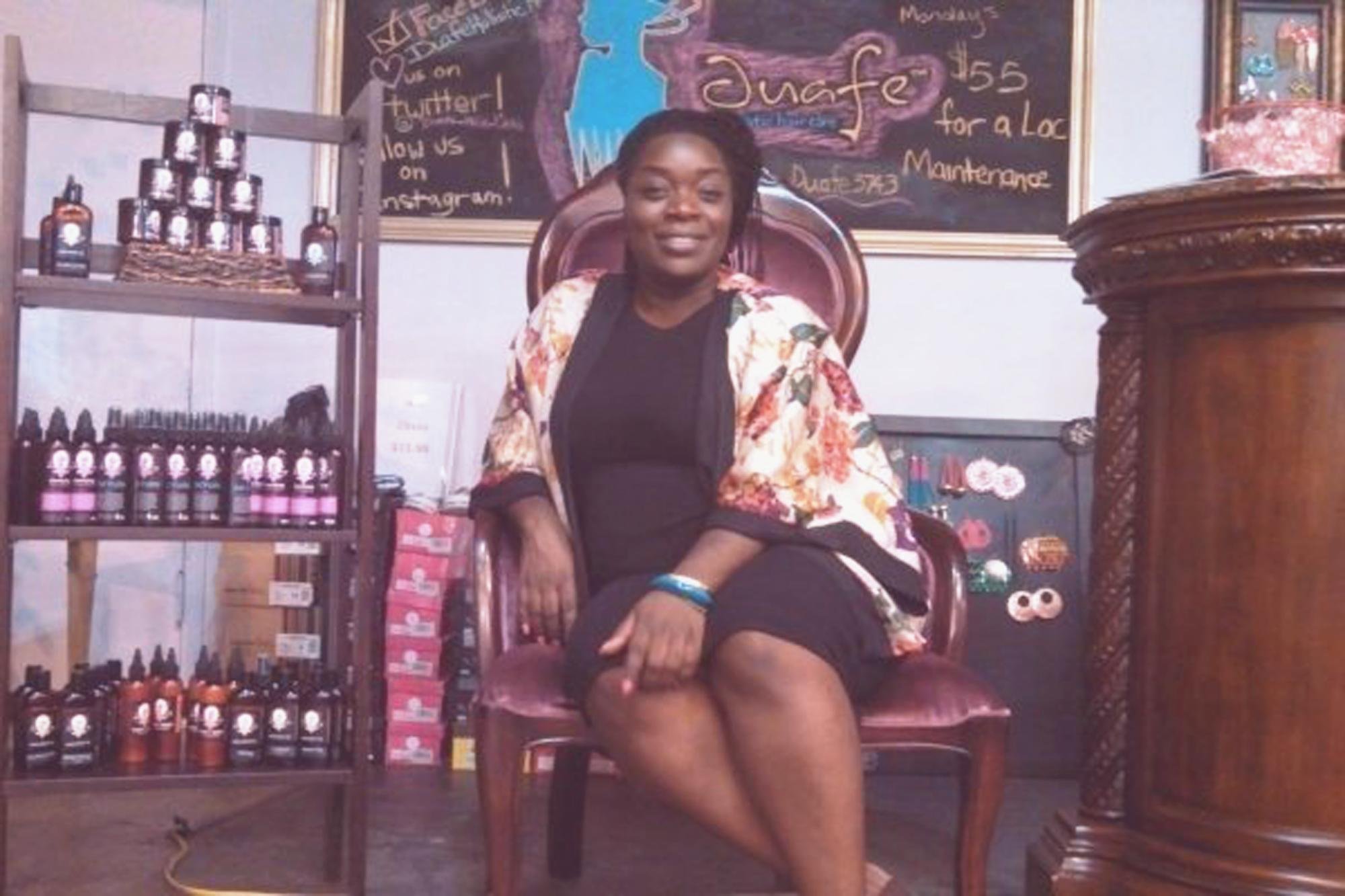 A woman with dark hair sitting in a vintage wooden chair inside a shop or boutique, surrounded by shelves of nail polish bottles and a chalkboard sign behind her advertising a manicure service.