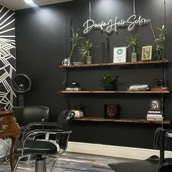Interior of a salon with black walls, wooden shelves holding potted plants, framed pictures, and hair products, with salon chairs in the foreground.