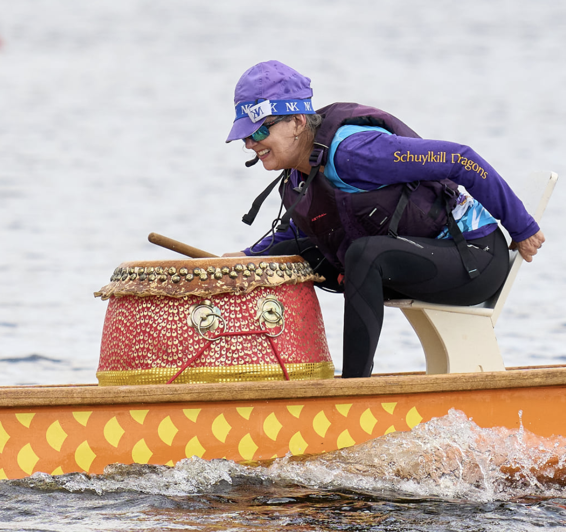 A woman wearing a purple and blue jacket, black leggings, and a purple cap, smiling as she paddles a dragon boat with a decorative drum mounted in front of her on the boat.