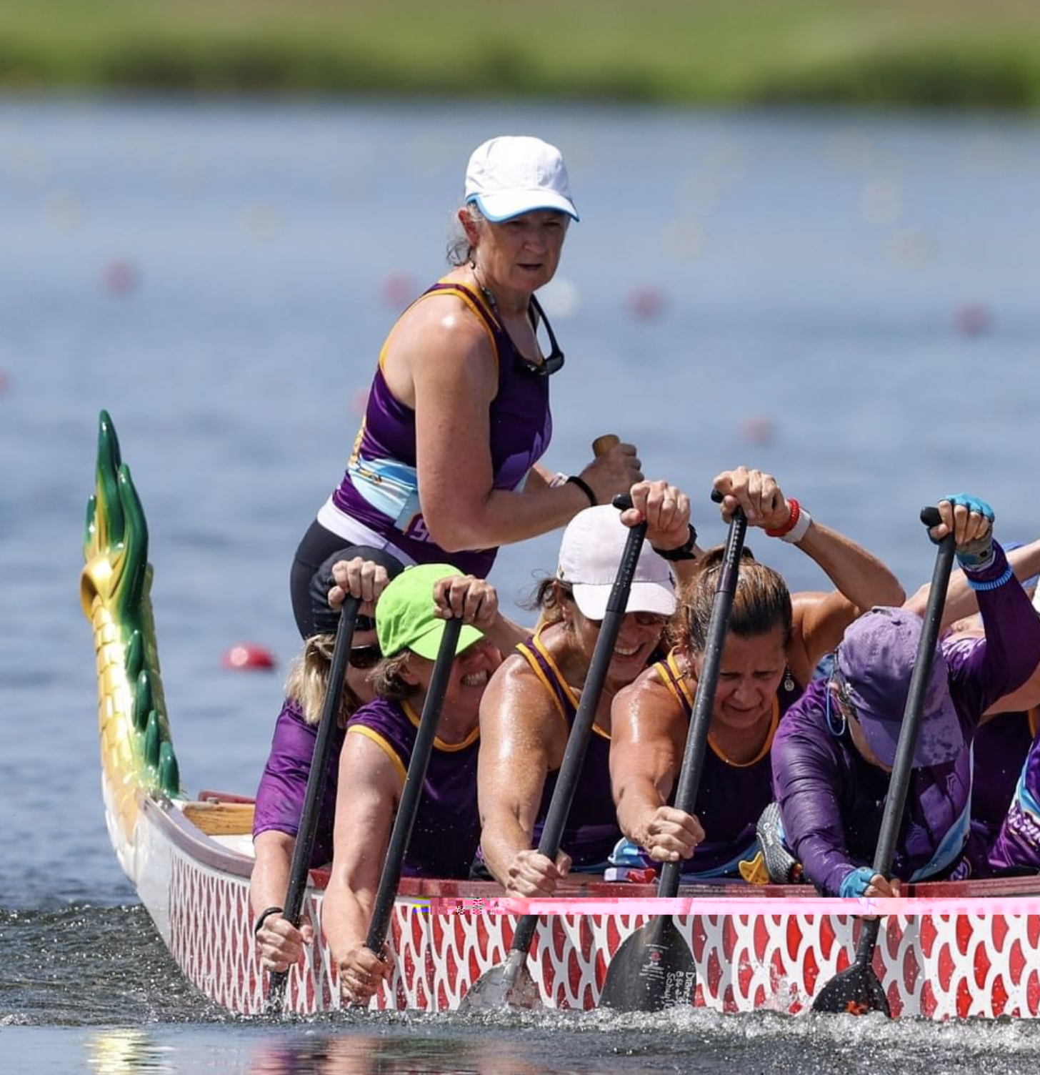 A group of women participating in a dragon boat race, wearing purple and yellow uniforms, paddling on the water with a purple and yellow dragon boat, one woman stands at the front wearing a white cap and holding a paddle.