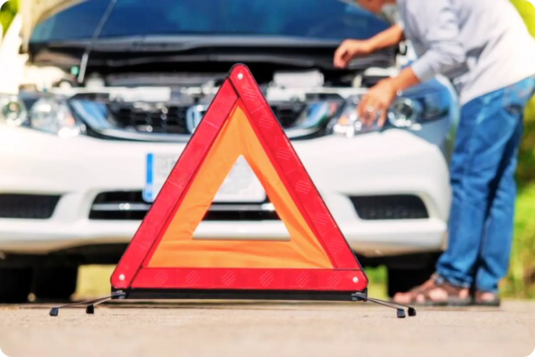 A warning triangle placed on the ground in front of a car with its hood open, and a person working on the engine.