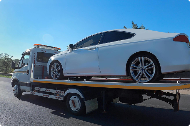 A white car is on a flatbed tow truck driving on a highway under a clear blue sky.