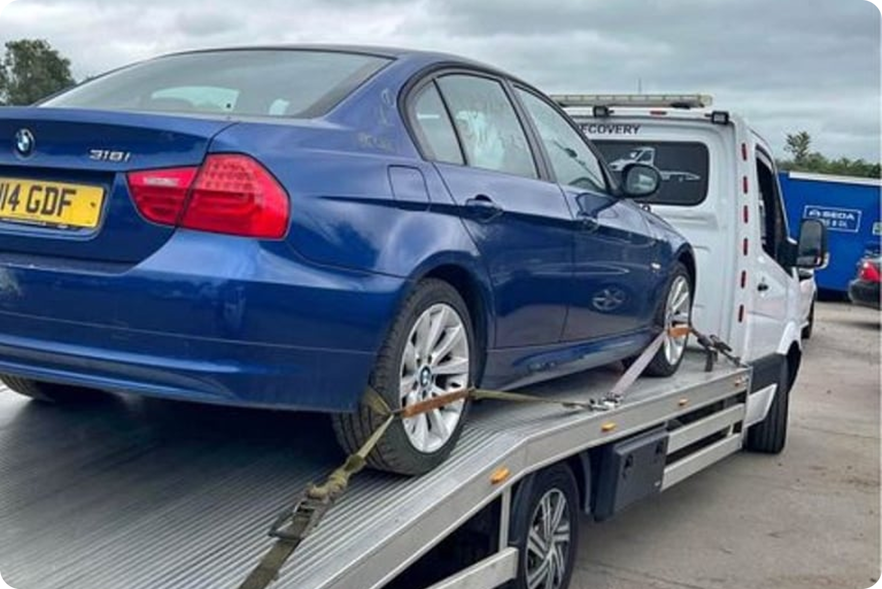 Blue BMW 3 Series sedan being transported on a flatbed tow truck, secured with straps.