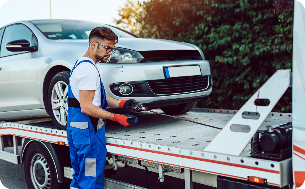 A mechanic in blue coveralls with orange gloves is working on a silver car that is loaded onto a flatbed tow truck, using a device attached to a cable.