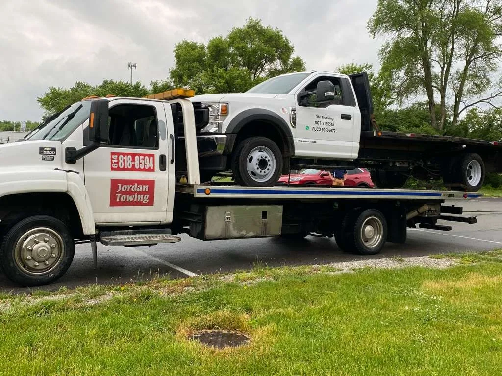 A flatbed tow truck transporting a white pickup truck with black accents and a black truck bed. The tow truck has signage for Jordan Towing and its phone number, with a background of green trees and an overcast sky.